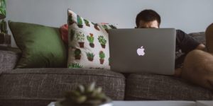 Man's face obscured by the laptop he is using on a grey living room couch.